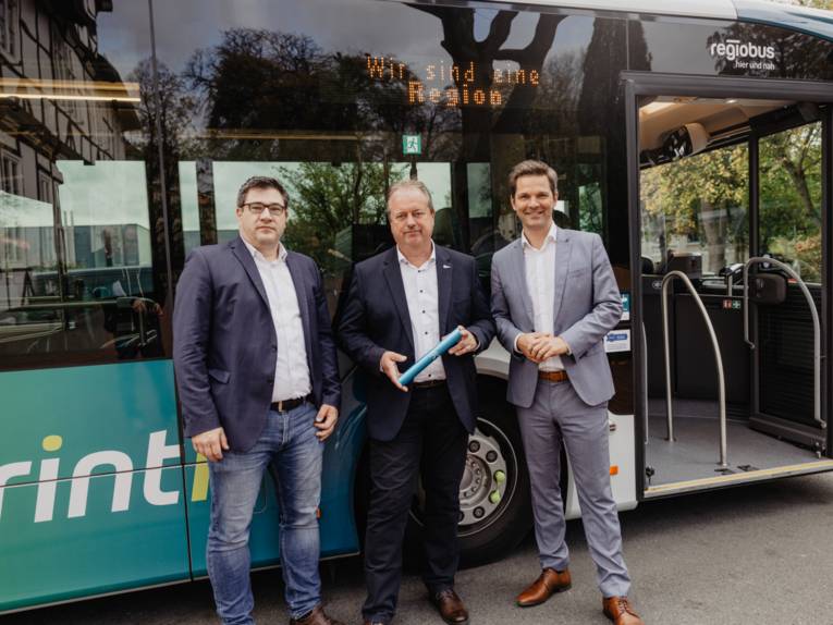 Drei Herren mit einem Staffelstab vor einem Bus